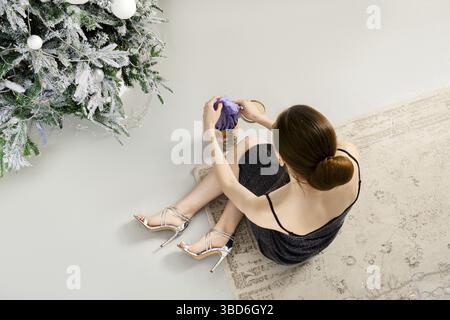 View from above at a woman in black glitter dress sitting on the floor near a Christmas tree and taking violet silk scarf out of a gift box Stock Photo