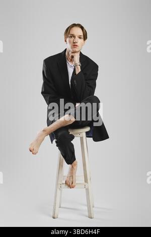 Young barefoot man in black suit sitting on tall chair in studio Stock Photo