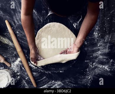 Person stretches dough on a dark surface dusted with flour. Stock Photo