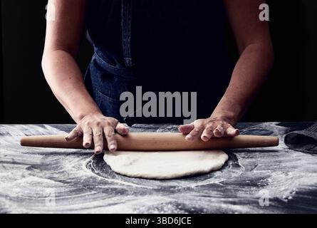 Person stretches dough on a dark surface dusted with flour. Stock Photo