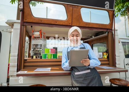 Indonesian southeast asian chinese muslim female barista in hijab standing while using a tablet in front of her coffee shop. A small business of a cof Stock Photo