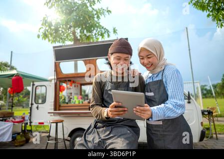 Indonesian southeast asian male and a Chinese muslim female in hijab couple baristas using a tablet in front of their coffee shop. A small business of Stock Photo