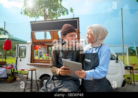 Indonesian southeast asian male and a Chinese muslim female in hijab couple baristas using a tablet in front of their coffee shop. A small business of Stock Photo