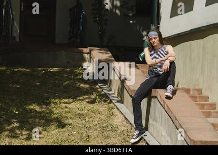 Creative long-haired man in tank top and jeans sits on railing Stock Photo