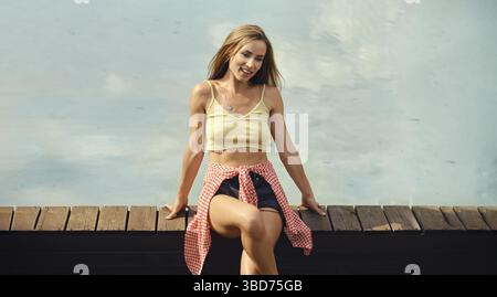 Positive smiling young woman sits on wooden railing of the bridge Stock Photo