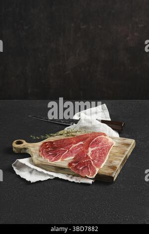 Thin slices of jamon iberico on a wooden cutting board, accompanied by a knife and a sprig of thyme. A white cloth adds a rustic touch to the dark, te Stock Photo