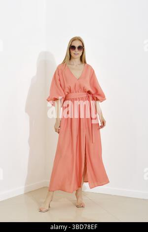 A full-length portrait of a woman in a coral maxi dress with a v-neckline and short sleeves, cinched at the waist, standing against white wall Stock Photo
