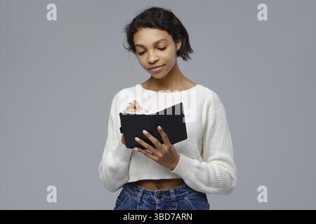 Concentrated young african woman standing against grey background and writing in diary Stock Photo