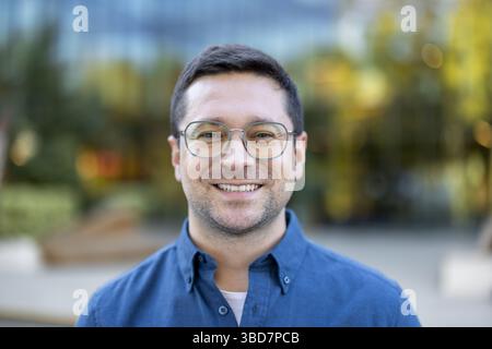 A portrait of a smiling man wearing glasses outdoors. He's dressed in a blue button-down shirt. A warm, inviting expression. Stock Photo