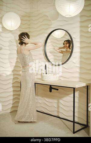 A woman dressed in a sparkling silver dress stands at a mirror, checking her reflection while delicately fixing her hair. Soft light emanates from han Stock Photo