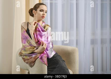 Cute young woman with red lips and natural makeup sits in armchair with her shoulders wrapped in colorful silk shawl Stock Photo