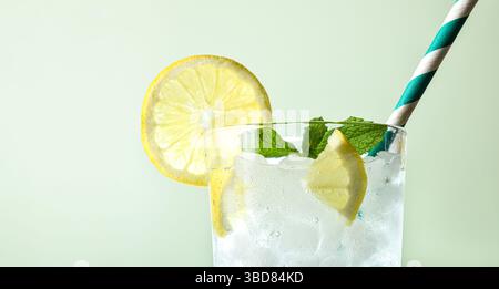 Very cold lemon soda drink with ice cubes in a glass cup decorated with mint leaves and a straw with a green isolated background. Front view. Stock Photo