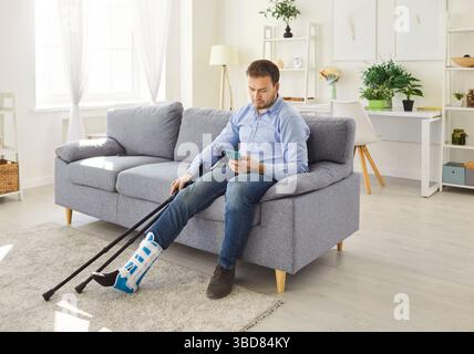 Male person with injured leg sitting on sofa at home using mobile phone. Stock Photo