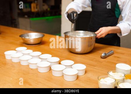Chef whisking cream mixture in a metal bowl with multiple empty white ramekins arranged on a wooden kitchen counter. Stock Photo