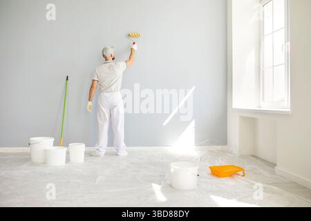 Male painter in uniform painting grey wall with roller in bright room during house construction. Stock Photo