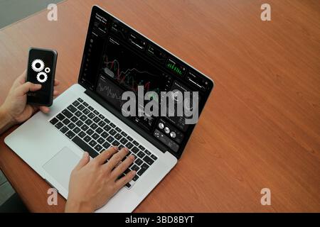 Close-up view of hands using a laptop and smartphone with digital interface displaying financial trading data and automation icons. Stock Photo
