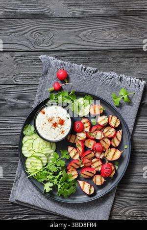 Grilled radishes with cucumber slices and sour cream sauce served on a dark grey plate on a black wooden table, vertical view, free space, real photo, Stock Photo