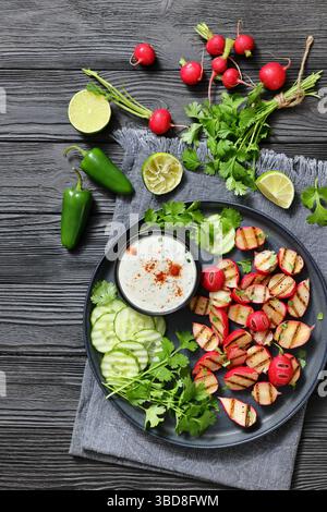 Grilled radishes with cucumber slices and sour cream sauce served on a dark grey plate on a black wooden table with ingredients and lime, vertical vie Stock Photo