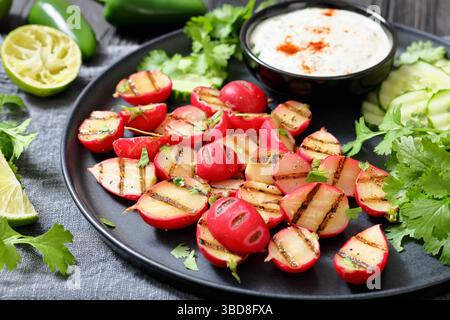 Grilled radishes with cucumber slices and sour cream sauce served on a dark grey plate, horizontal view, close-up, real photo, not AI generated Stock Photo