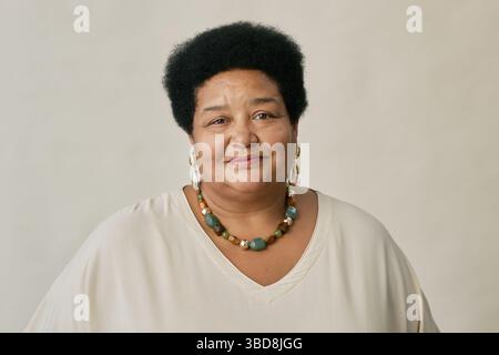 Portrait of mature African American woman with short curly hair wearing beaded necklace and white top, smiling softly at camera in studio Stock Photo