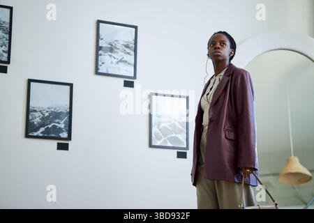 Portrait of an African American woman wearing a blazer and carrying a handbag while observing art pieces in a modern gallery setting Stock Photo