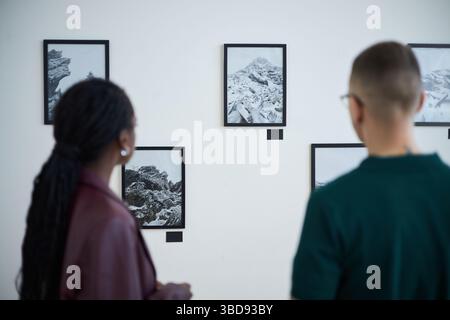 Individuals observing framed artwork on gallery wall during exhibition. Black and white landscape photographs displayed with intriguing compositions a Stock Photo
