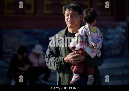 Smiling man in green jacket carrying a toddler dressed in colorful patterned clothes in a temple courtyard Stock Photo