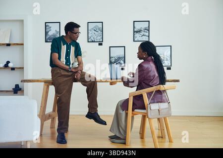 African American couple collaborating on work project in modern art gallery. Seated woman using laptop while man standing beside table, discussing pro Stock Photo