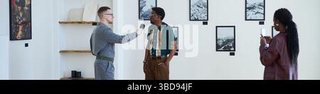 Header with black man standing in an art gallery, being interviewed by a man holding microphone while woman taking notes is nearby. Displayed black an Stock Photo