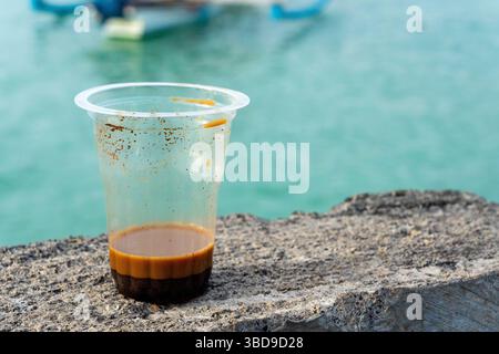 Plastic cup with coffee in it sits on wall on the beach. The cup is almost empty. The scene is peaceful and serene, with the water Stock Photo