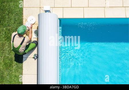 A professional is checking the pool water quality beside a sparkling blue pool in a residential backyard. The grass is lush and green, indicating a wa Stock Photo