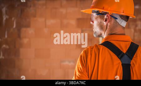 A construction worker in an orange uniform and hard hat examines the progress of a building project. The individual stands against a textured wall, fo Stock Photo