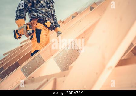 A construction worker stands atop a roof, wearing a plaid shirt and protective gear. They are focused on using a power tool to secure wooden beams. Stock Photo