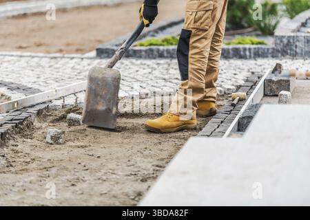 A construction worker in sturdy gear is carefully placing stone pavers into a garden pathway. The project takes place in a residential area under clea Stock Photo
