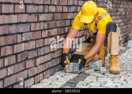 A worker kneels near a brick wall, carefully aligning and placing stones on the ground. The sun shines brightly, highlighting the construction activit Stock Photo