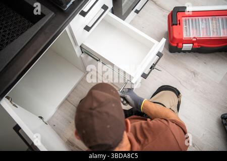 A worker is installing a kitchen drawer in a stylish modern cabinet. Tools are nearby, and the floor features light-colored planks. Attention to detai Stock Photo