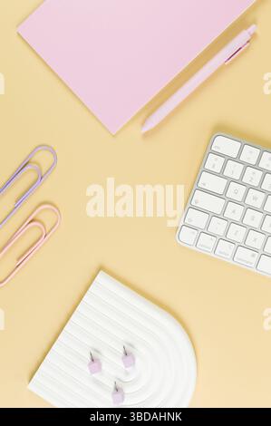 Top view of white keyboard, pink notebook, pink pen on yellow background. Paper clips, pins, modern office, school. White arch tray, flat lay. Stock Photo