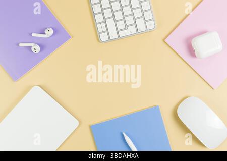 Top view of white keyboard, mouse, touchpad, earphones case, pen on yellow background. Purple, pink and blue notebooks, modern office, school. Stock Photo