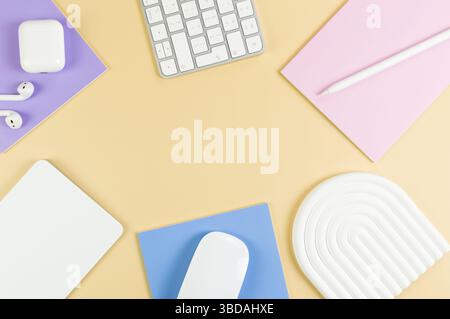 Top view of white keyboard, mouse, touchpad, earphones case, pen on yellow background. Purple, pink and blue notebooks, modern office, school. Stock Photo