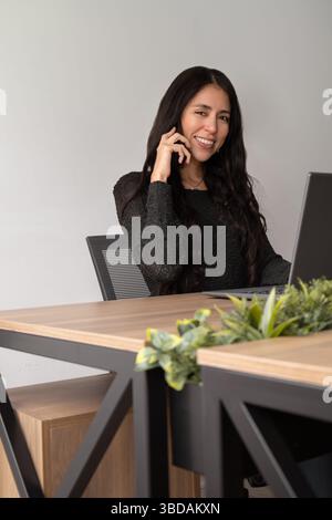 A young professional woman speaks on the phone while working in a modern office, demonstrating multitasking and effective communication in a business Stock Photo