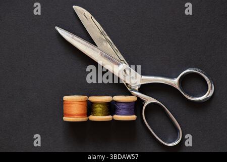 Dressmaker scissors and wooden spools of thread on black background, sewing and cutting tools Stock Photo