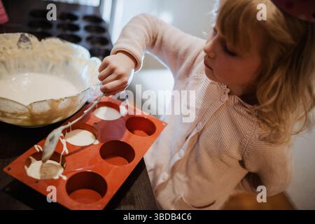 Two siblings are joyfully baking together in a warm kitchen. One sibling carefully fills silicone molds with batter while another watches, creating sw Stock Photo