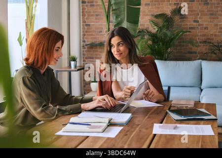 Two happy diverse business women working using laptop at desk in green office. Stock Photo