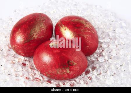 Three red-coloured, partially gold-plated Easter eggs in a nest made of colourless plastic stones Stock Photo