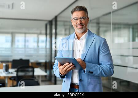 Confident mature business man in suit standing in office holding tab, portrait. Stock Photo