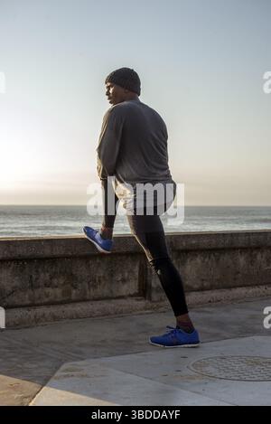 A man with exersise attire and a beanie on his head does leg stretches by the promonade as he views the ocean Stock Photo