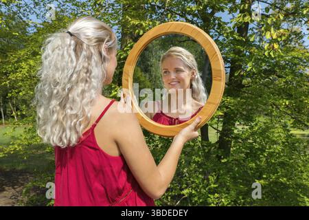 Young blonde caucasian woman looking in round wooden mirror outside Stock Photo