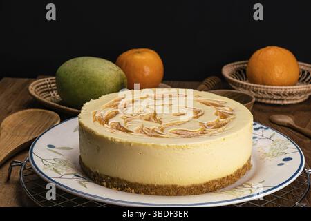 Side view of homemade mango cheesecake in ceramic dish on metal cooling rack with mango. ripe oranges, wooden spoon, cup and bamboo tray on wooden tab Stock Photo