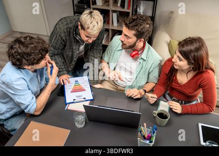 Top-down shot of multicultural young coworkers reviewing a colorful funnel chart and planning a digital marketing strategy around a laptop in a casual Stock Photo