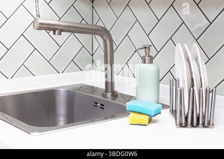 Stainless steel sink with clean plates in dish rack, soap bottle, and sponges on a clean countertop against herringbone tiles backsplash Stock Photo